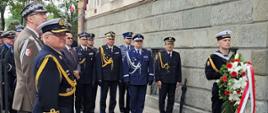 Przedstawiciele służb mundurowych oraz wojska stoją na baczność przed nimi stoi żołnierz z wiązanką kwiatów.