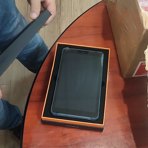 Ręka osoby wyjmującej tablet z czarno-pomarańczowego pudełka, obok otwartego kartonu z folią bąbelkową na drewnianym stole.