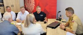 Kilka osób siedzi przy stole konferencyjnym podczas spotkania w siedzibie Państwowej Straży Pożarnej. Po prawej stronie strażak w mundurze rozmawia z uczestnikami, a w tle widoczna jest czerwona ścianka z godłem Polski i logo Państwowej Straży Pożarnej.