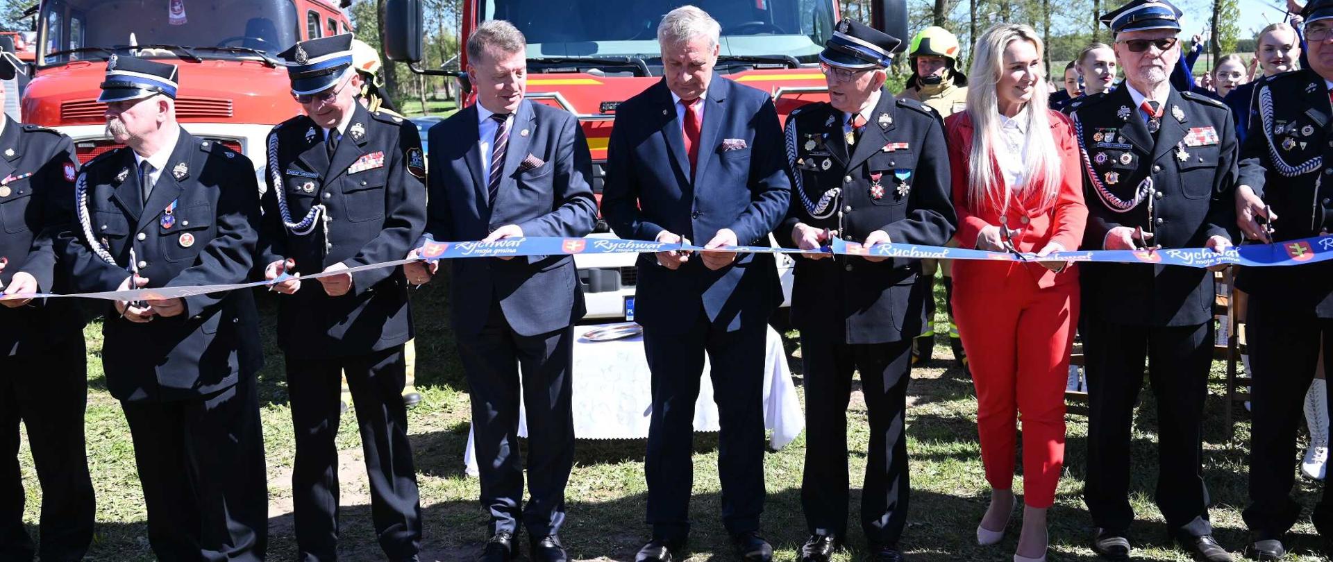 Obchody Dnia Strażaka w Rozalinie. Goście przecinają wstęgę nożyczkami. W tyłu samochody strażackie 