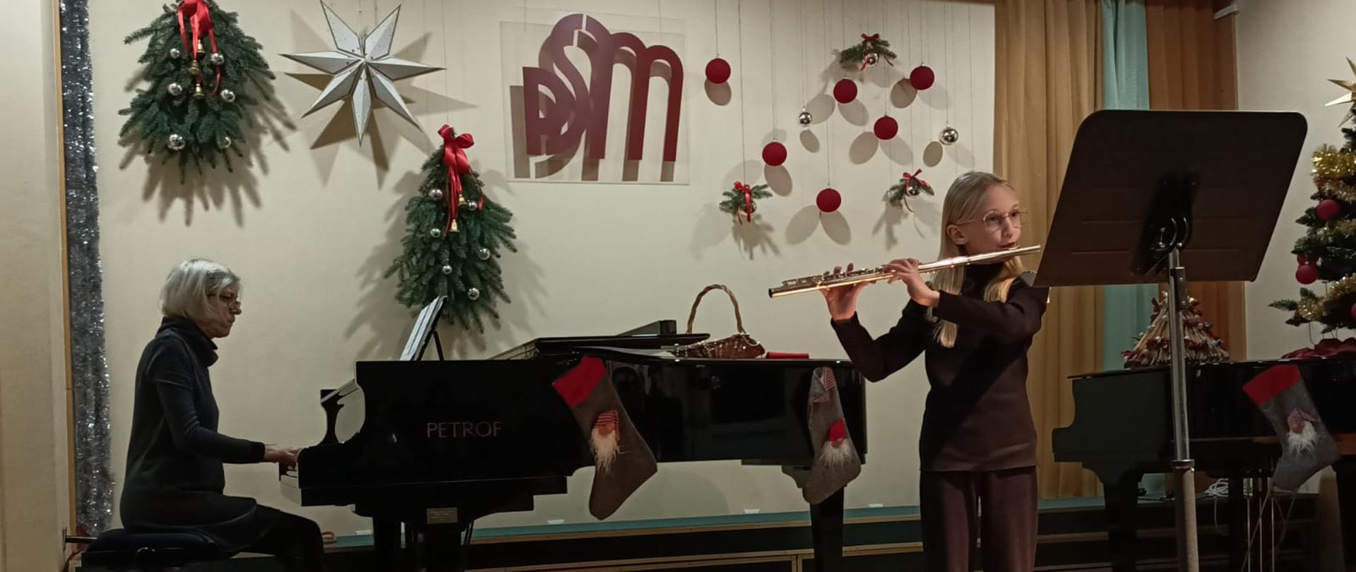 zdjęcie - grająca na flecie uczennica, za nią fortepian przy fortepianie Pani nauczycielka akompaniuje uczennicy, foto. L. Zegadło-Kołda