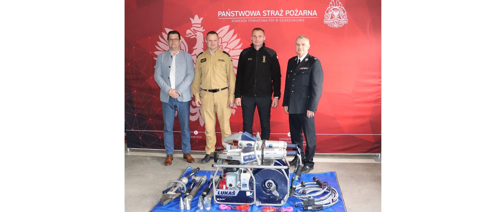 Trzech strażaków i burmistrz stoją przed czerwonym tłem z napisem Państwowa Straż Pożarna – Komenda Powiatowa PSP w Dzierżoniowie. Na podłodze leży niebieska mata, a na niej rozłożony zestaw hydraulicznych narzędzi ratowniczych LUKAS, w tym pompa, nożyce, rozpieracz i akcesoria. Sprzęt wygląda na prezentowany podczas oficjalnego przekazania