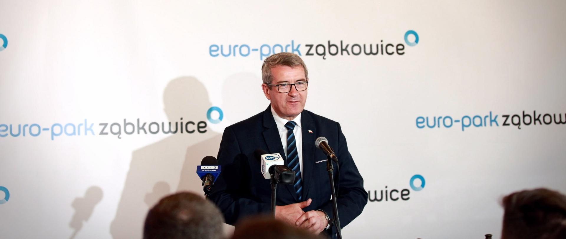 Minister przemawia do mikrofonu na tle białej ściany z napisami Euro-park Ząbkowice.
