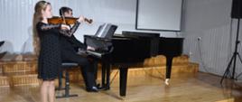 Na zdjęciu wykonanym w auli PSM widnieje uczennica grająca na skrzypcach z akompaniamentem fortepianu. Przy fortepianie nauczyciel Mateusz Gałuszka. Kolorystyka zdjęcia jest biało-czarno-brązowa.