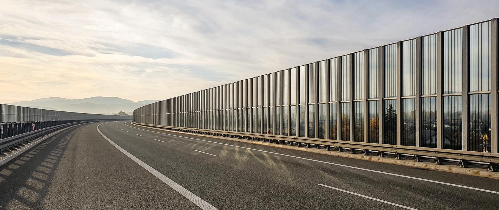 Widok na dwujezdniową drogę ekspresową z poziomu terenu. Droga przebiega w łuku poziomym. Na widnokręgu widoczne są góry, niebo jest zachmurzone. Drogę wyposażono w przezierne ekrany akustyczne oraz bariery ochronne.