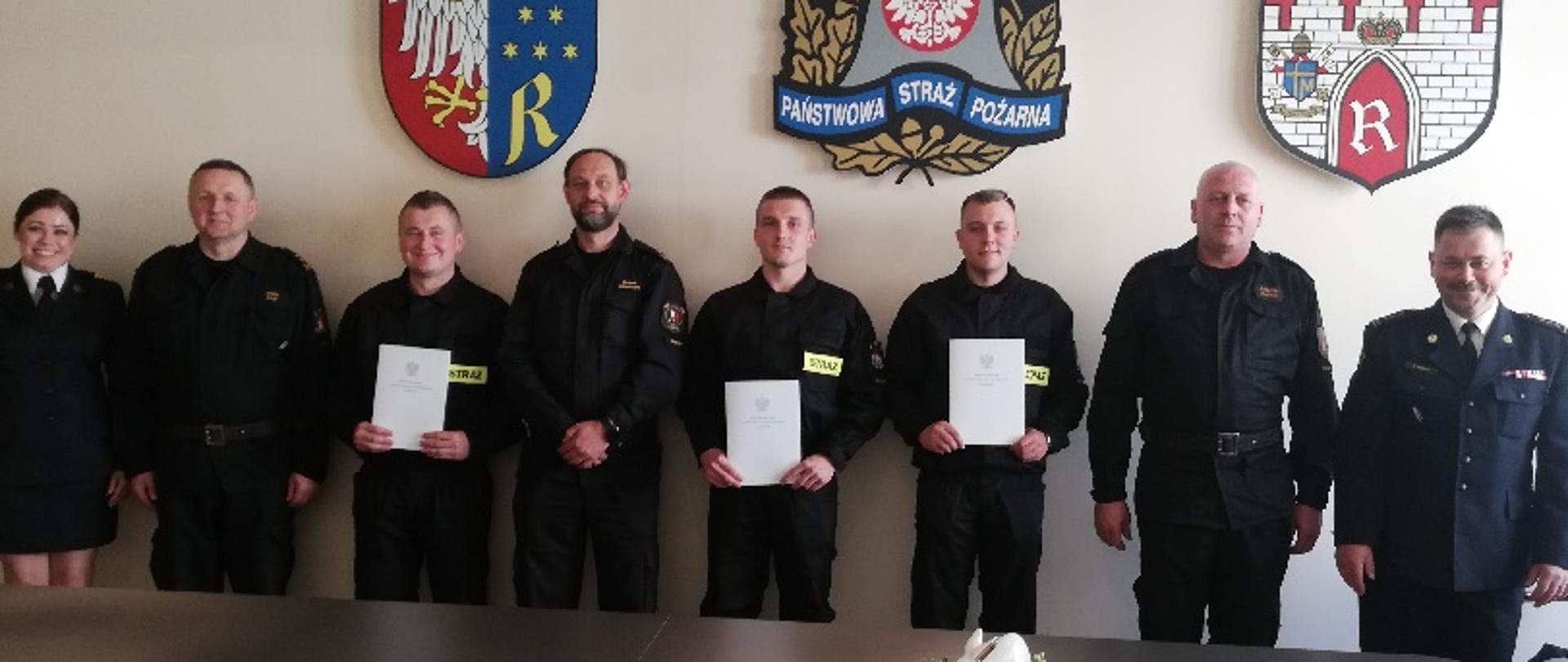 Na zdjęciu widać pełniącego obowiązki komendanta miejskiego Państwowej Straży Pożarnej w Radomiu st. bryg. Marcina Ziomka w asyście dowódców jednostek ratowniczo – gaśniczych oraz funkcjonariuszki wydziału kadr komendy miejskiej. Komendant oraz funkcjonariuszka ubrani są w mundury galowe. Kadra dowódcza w ubrania dowódczo - sztabowe w kolorze czarnym. Pomiędzy nimi stoją trzej nowo przyjęci do służby funkcjonariuszy. Strażacy ustawieni są w rzędzie jeden obok drugiego. W rękach trzymają akty przyjęcia do służby. Ubrani są w czarne ubrania koszarowe z jaskrawymi elementami odblaskowymi. Zdjęcie wykona w sali konferencyjnej Komendy Miejskiej Państwowej Straży Pożarnej w Radomiu. 