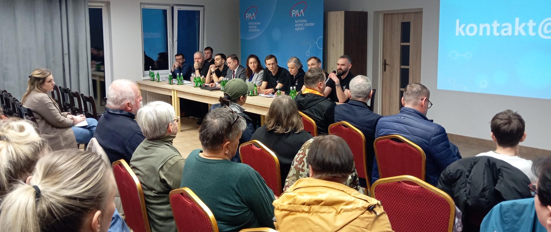 Spotkanie ekspertów PAA z mieszkańcami gm. Choczewo - eksperci siedzą za stołem, jeden przemawia do mikrofonu. Na przeciwko grupa mieszkańców