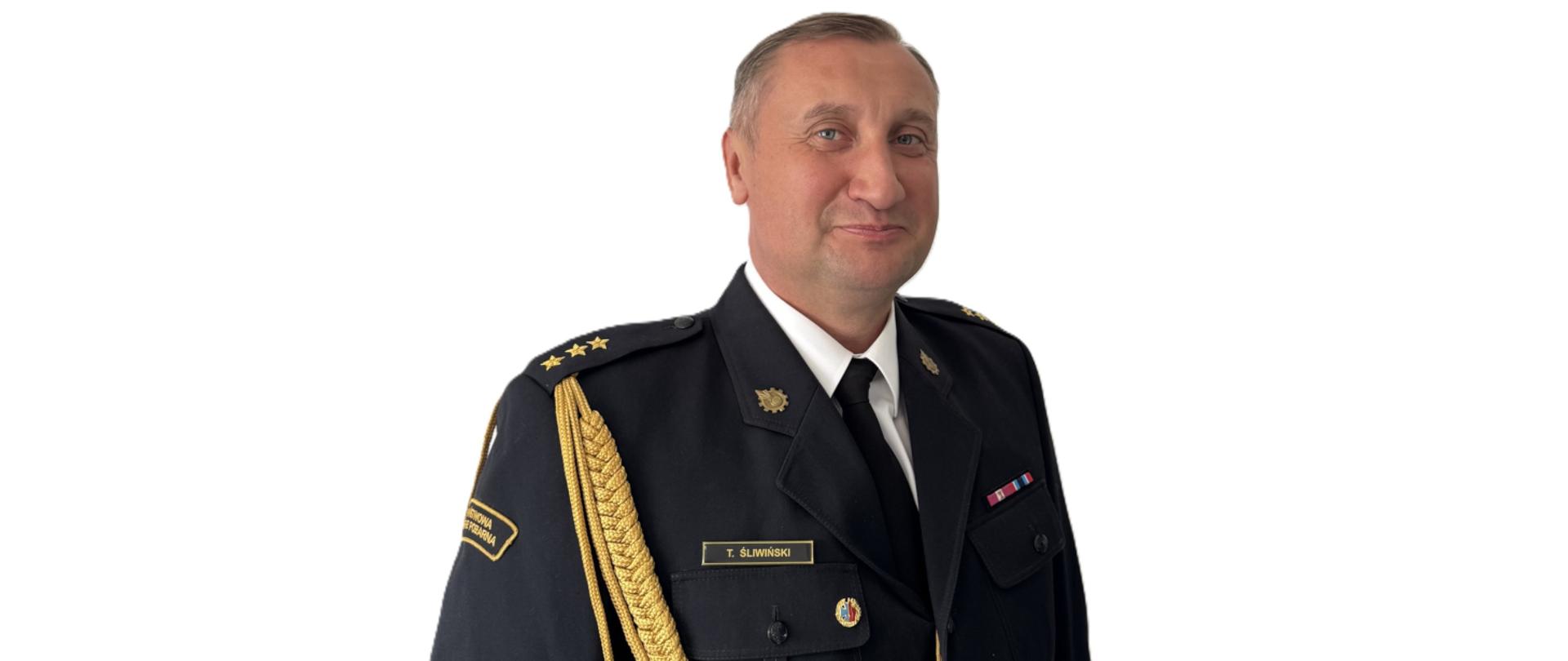 Zdjęcie przedstawia mężczyznę w formalnym mundurze służbowym. Ma krótkie, jasne włosy i poważny wyraz twarzy. Mundur jest granatowy, z krawatem, ozdobnymi złotymi sznurami na ramieniu oraz odznaczeniami i naszywkami na piersi. Tło jest jasne i jednolite.