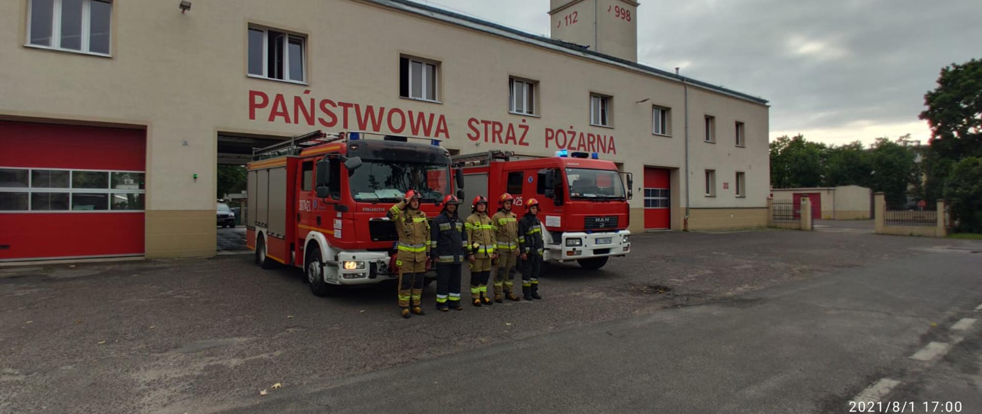 Strażacy stojący na baczność w tle samochody pożarnicze oraz budynek jednostki