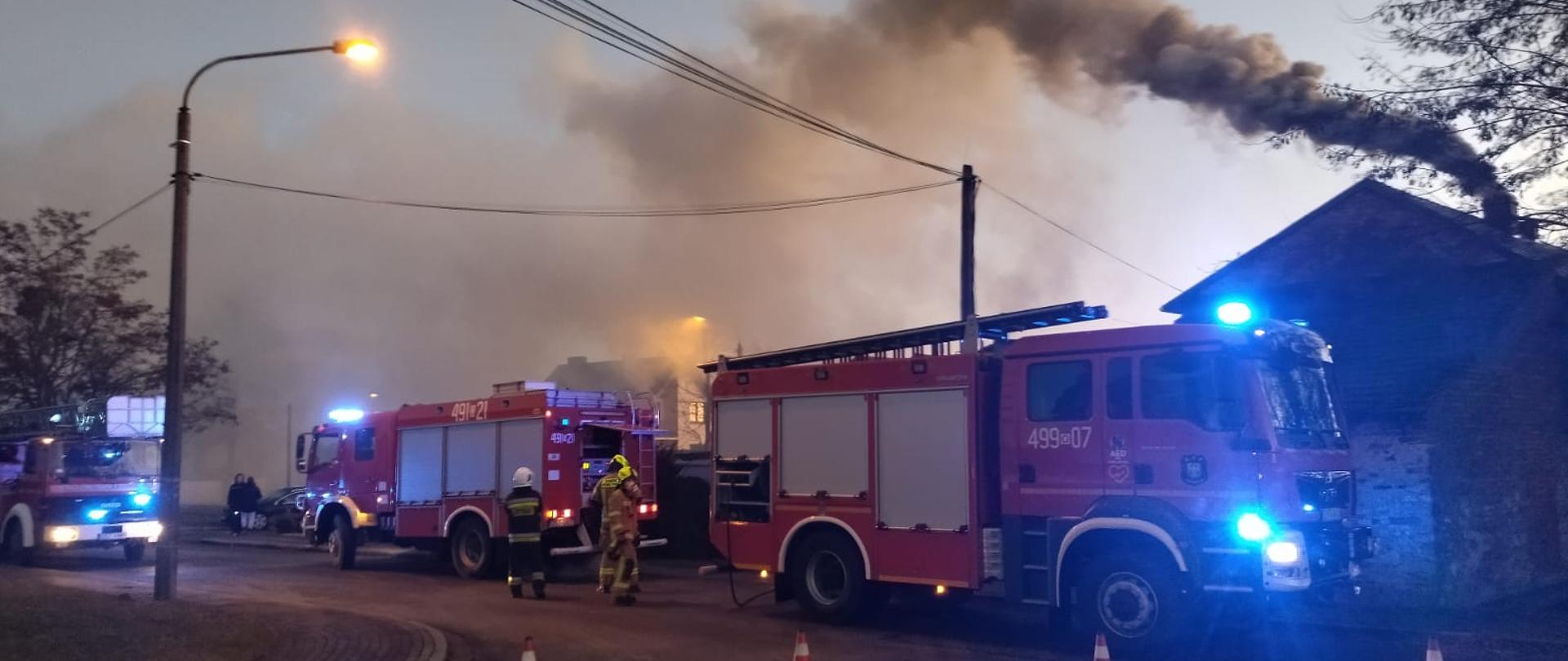 Zdjęcie przedstawia dwa samochody pożarnicze na tle budynku z dymiącym kominem