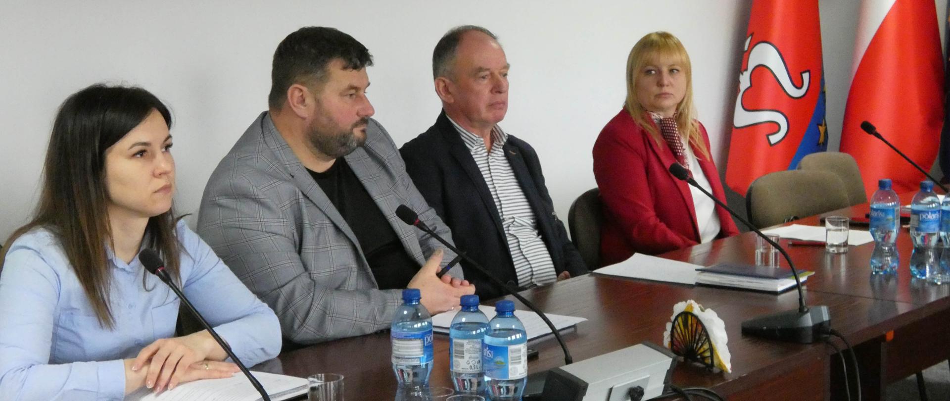 Cztery osoby siedzące przed brązowym stołem. Dwie kobiety i dwóch mężczyzn. Przed nimi są mikrofony. Ściany w pomieszczeniu są jasne. W tle widać stojące flagi w rogu pokoju. Na stole leżą butelki z wodą oraz szklanki. 