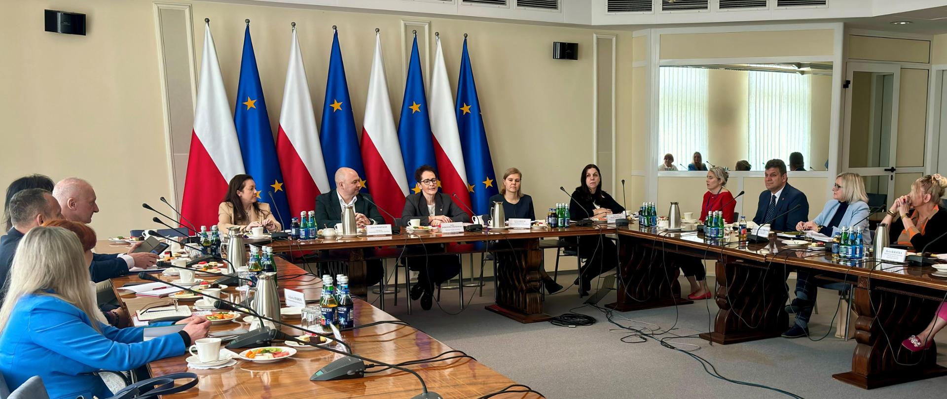 Szefowej służby cywilnej rozmawia przy konferencyjnym stole ze związkami zawodowymi, za nimi flagi Polski i Unii Europejskiej