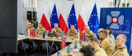 Uczestnicy międzynarodowego spotkania PSP siedzą przy dużym stole konferencyjnym. W tle szereg flag: polskie, unijne i norweskie. Na stole woda, owoce i materiały szkoleniowe.
