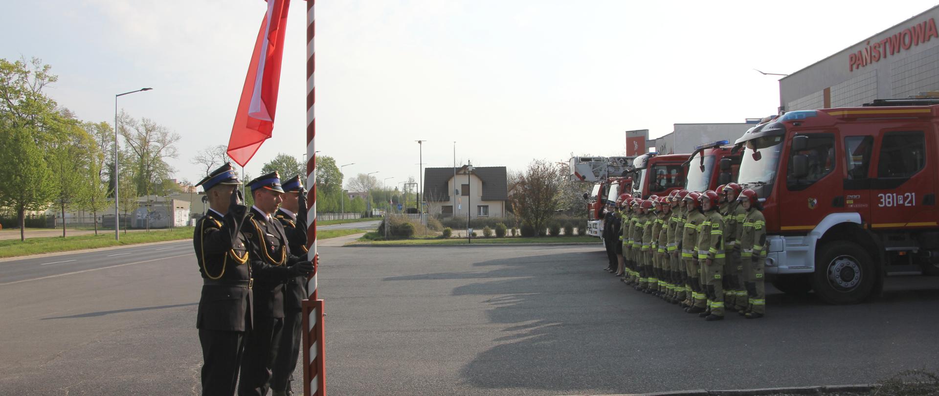Strażacy stoją na baczność przy maszcie. W tle w szeregu stoją strażacy na tle samochodów i komendy.