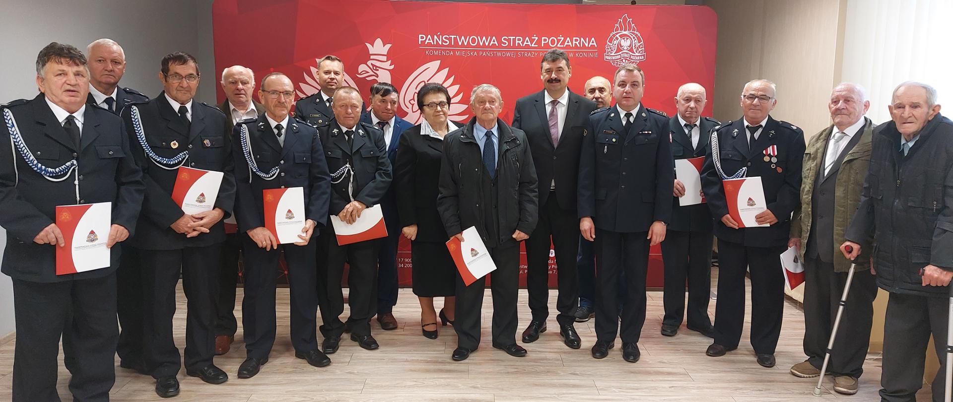 Zdjęcie przedstawia strażaków oraz druhów OSP, którzy otrzymali od komendanta decyzje o przyznaniu świadczenia ratowniczego, Strażacy oraz druhowie stoją przed banerem komendy. 