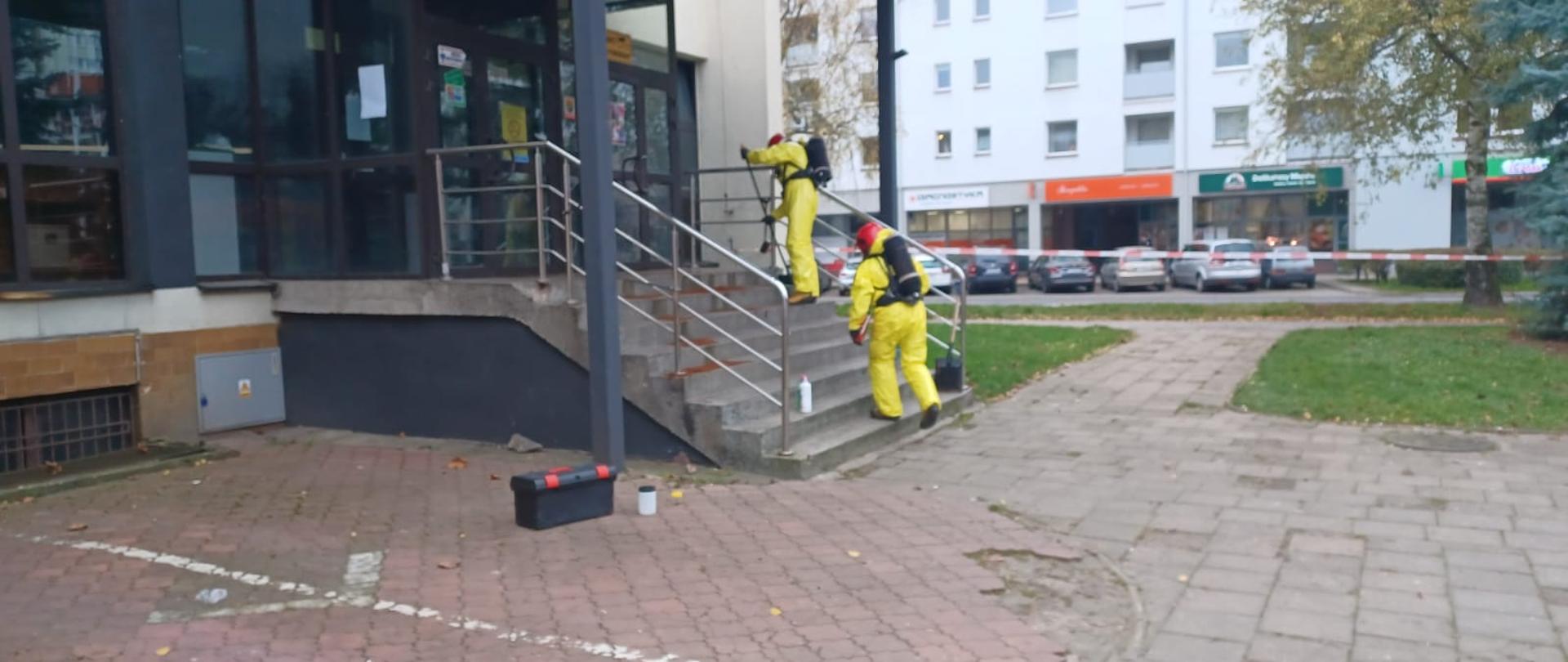 Na zdjęciu widać dwóch strażaków zabezpieczonych w ubrania przeciwchemiczne którzy pobierają próbkę substancji.