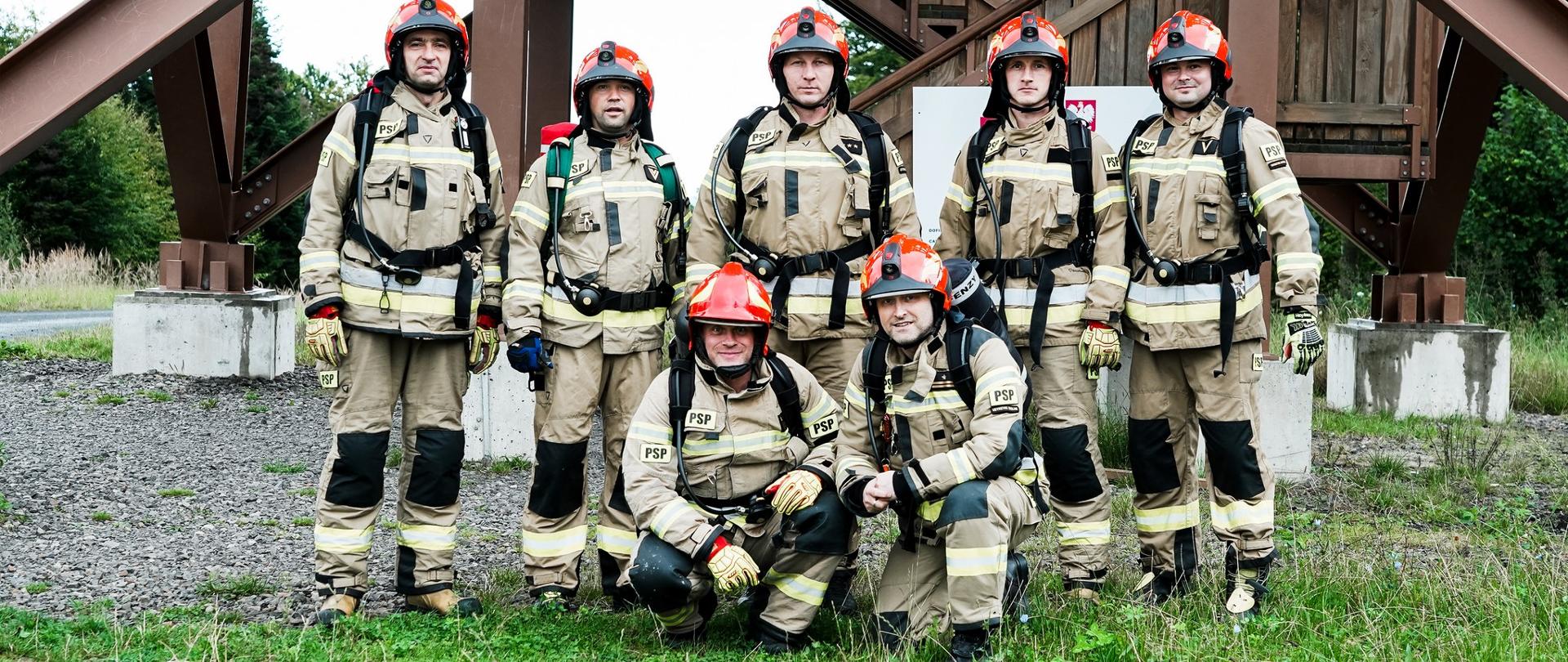 Zdjęcie przedstawia strażaków PSP z Ustrzyk Dolnych w ubraniach specjalnych pod wieżą widokową na Holicy. 