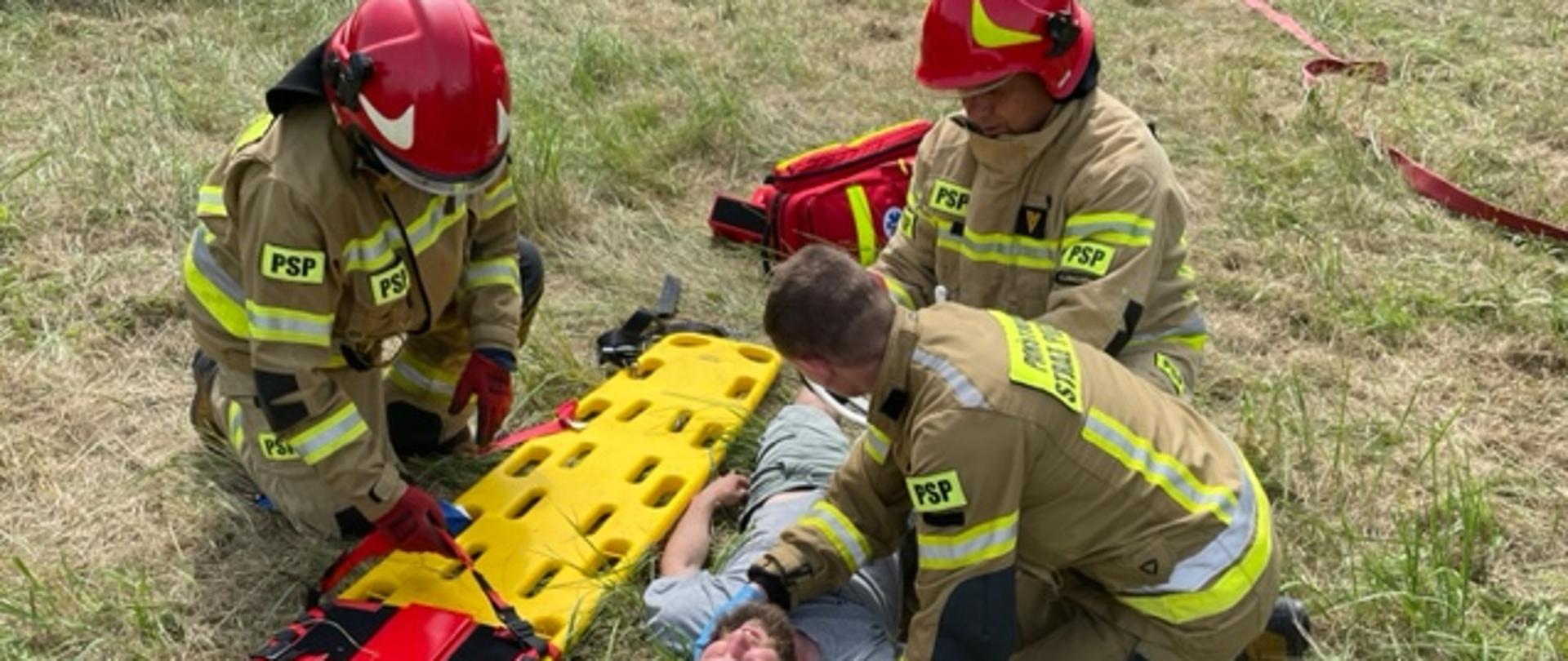 Zdjęcie przedstawia strażaków udzielających kwalifikowaną pierwsza pomoc osobie poszkodowanej. osoba leży na plecach a obok niej klęczy 3 strażaków w ubraniu specjalnym.