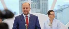 Pod półokrągłym oknem stoi wiceminister Lubnauer, przed nią dyrektor IBE w granatowym garniturze mówi do mikrofonu na stojaku.
