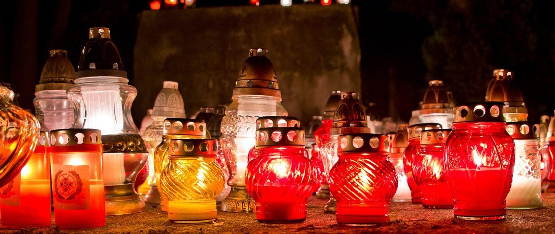 Zdjęcie przedstawia różnokolorowe zapalone znicze ustawione obok siebie na cmentarzu. Znicze różnych wielkości, w kolorach białym, czerwonym oraz żółtym. Zdjęcie wykonane w porze nocnej.