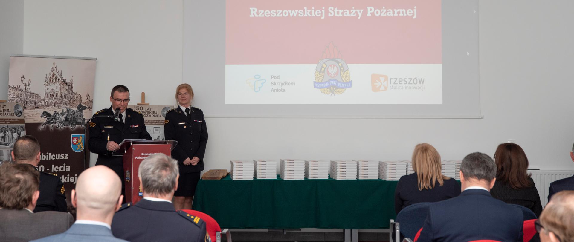 Zdjęcie przedstawia przemówienie komendanta miejskiego PSP w Rzeszowie st. bryg. Tomasza Barana stojącego przy mównicy. Na zdjęciu widoczni są słuchacze, prezentowane książki oraz na ścianie wyświetla się slajd "150 lat Rzeszowskiej Straży Pożarnej"