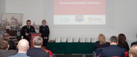 Zdjęcie przedstawia przemówienie komendanta miejskiego PSP w Rzeszowie st. bryg. Tomasza Barana stojącego przy mównicy. Na zdjęciu widoczni są słuchacze, prezentowane książki oraz na ścianie wyświetla się slajd "150 lat Rzeszowskiej Straży Pożarnej"