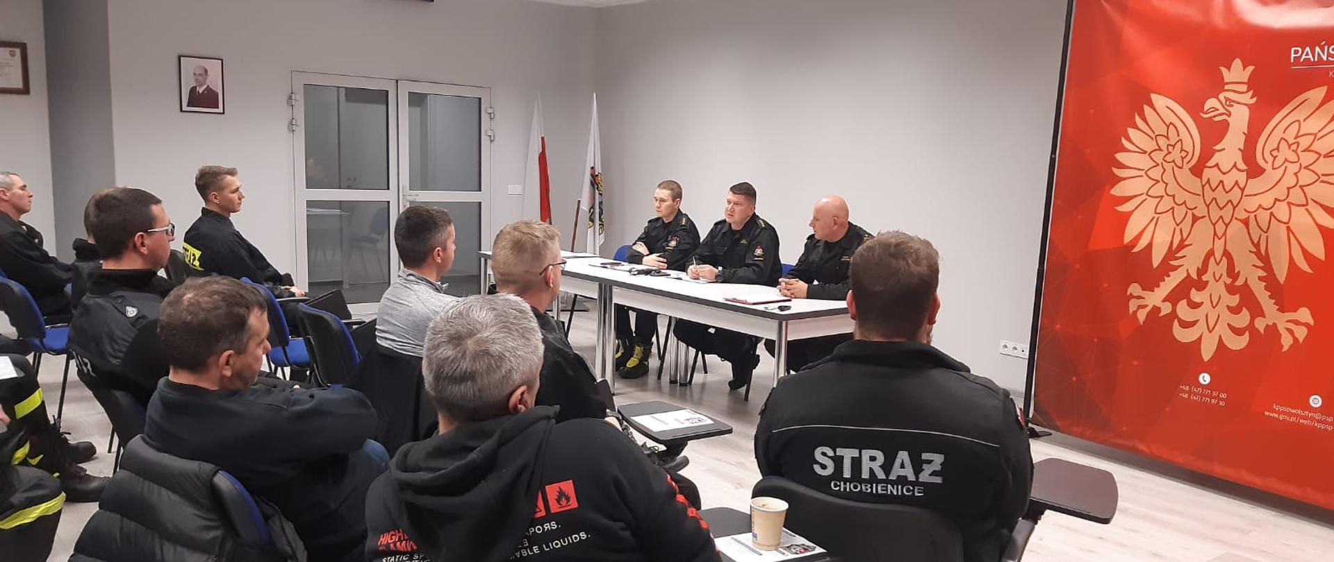 Zastępca Komendanta Powiatowego przemawia do zebranych druhów. Wszyscy siedzą na krzesłach. Po prawej stronie stoi baner PSP.