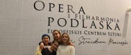 Zdjęcie przedstawia trzy uczennice i ich nauczycielkę, stojące na tle białej ściany z logo Opery i Filharmonii Podlaskiej - Europejskiego Centrum Sztuki im. S. Moniuszki.