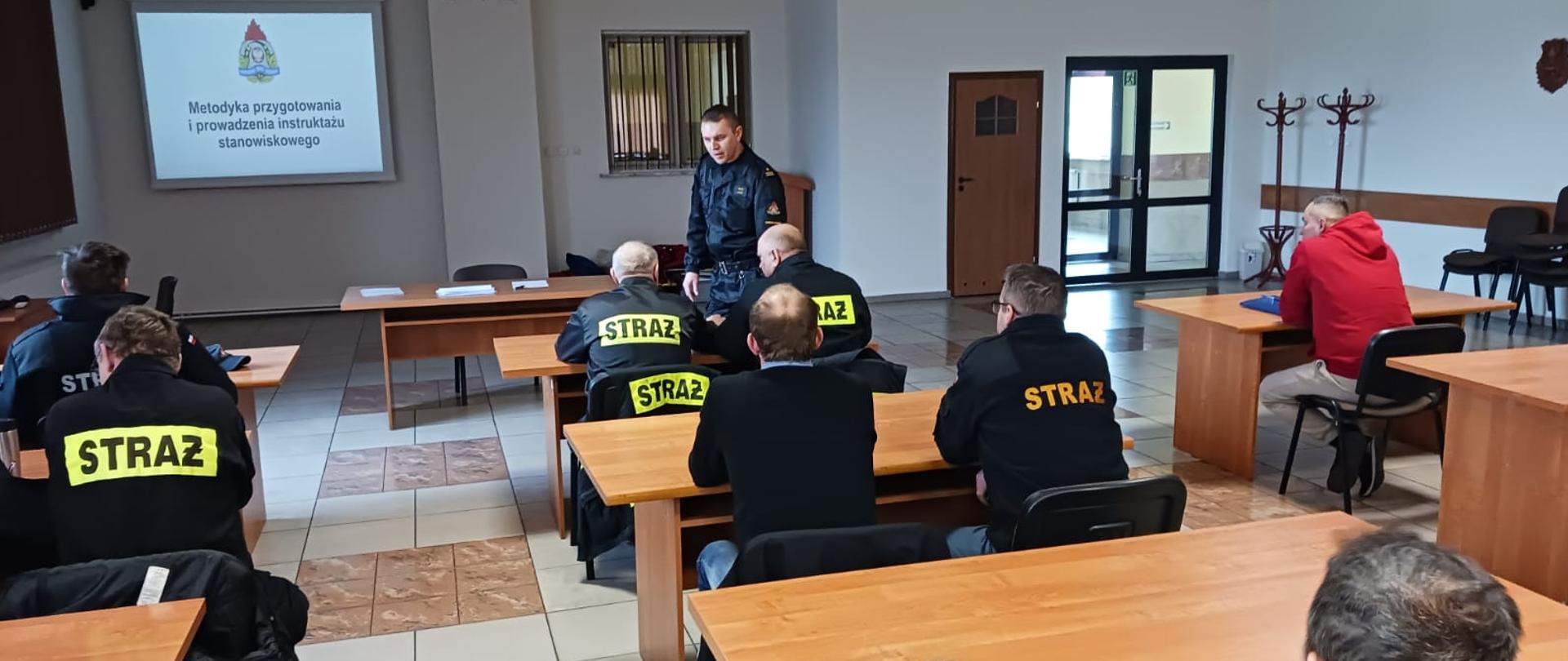 Zdjęcie przedstawia strażaka prowadzącego szkolenie z zakresu BHP oraz słuchających wykładu druhów OSP.