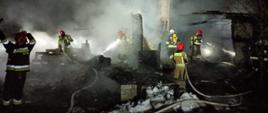 Strażacy pracujący przy pożarze pustostanu. Są ubrani w ubrania specjalne oraz aparaty ochrony układu oddechowego. Pogorzelisko jest spowite dymem.