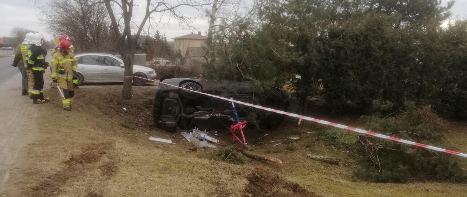 Na zdjęciu widać rozbity samochód osobowy, który znajduje się w pozycji na boku. Strażacy w celu stabilizacji pojazdu użyli podpory. Teren ogrodzony jest taśmą ostrzegawczą. Na zdjęciu widać grupę strażaków. Jeden z nich w ręku ma detektor napięć