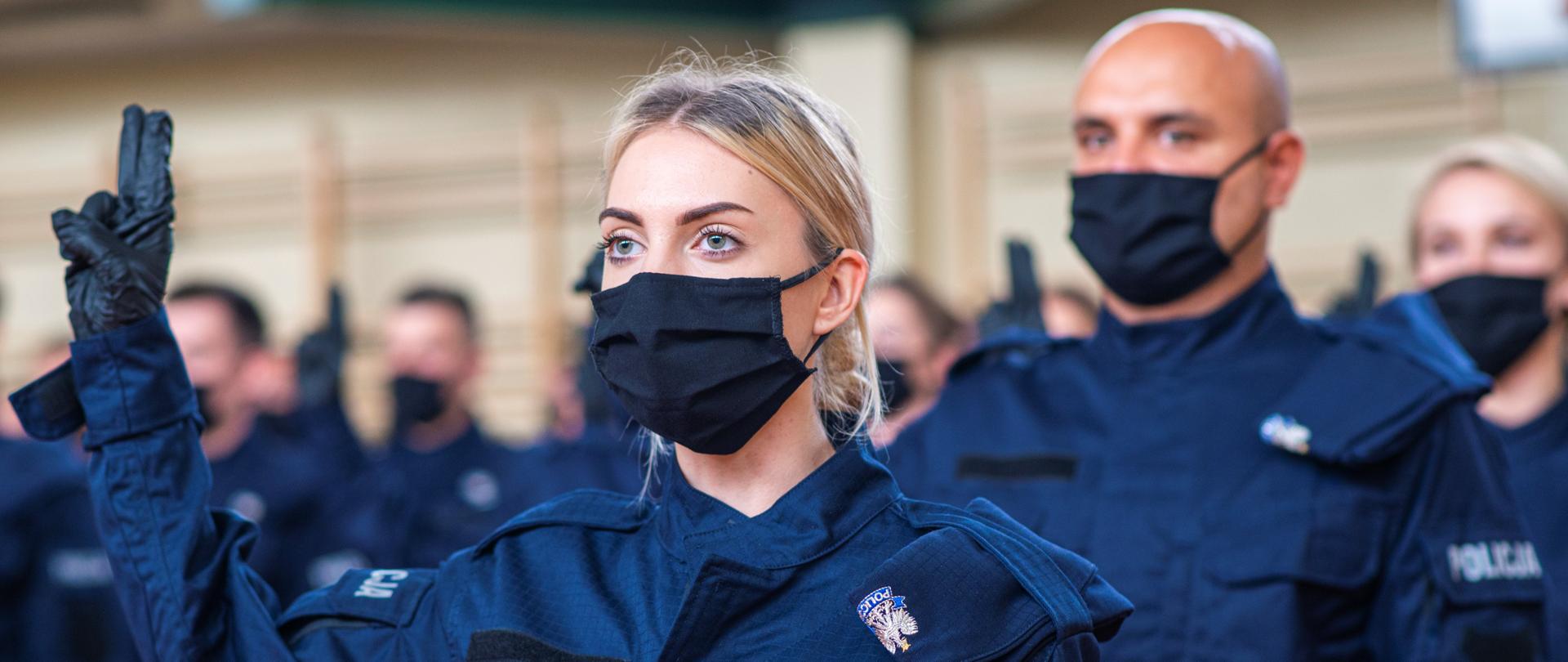 Policjanta ubrana w strój służbowy podnosząc dwa palce prawej dłoni i ślubuje w Oddziale Prewencji Policji w Łodzi, w tle widać innych ślubujących policjantów