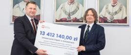 Minister Czarnek i mężczyzna w garniturze stoją i trzymają razem symboliczny czek z napisem 13 412 140 zł, za nimi na ścianie wiszą 3 portrety.