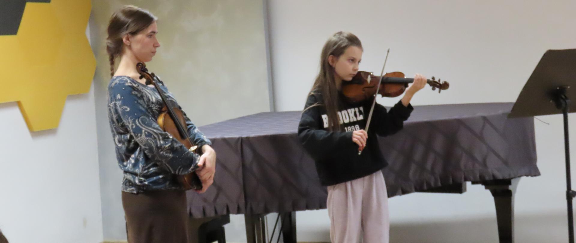 Z lewej strony prowadząca warsztaty trzyma w rękach skrzypce pośrodku dziewczynka gra na skrzypcach patrzy w stronę czarnego pulpitu z nutami który stoi przed nią.