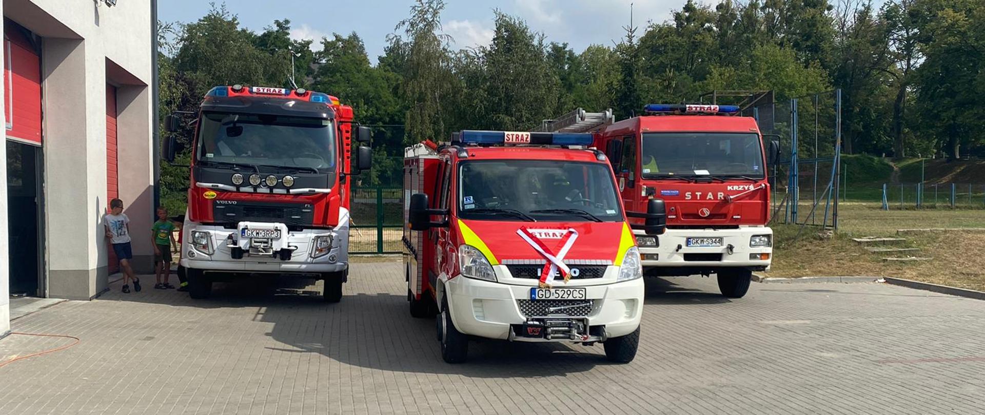 Nowy samochód gaśniczy ze wstążkami na przedniej masce stoi za nim cofnięte dwa samochody gaśnicze.