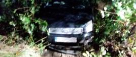 Przód samochodu po zderzeniu z innym pojazdem znajduje się w przydrożnym rowie. Widoczna jest tylko przednia część maski pojazdu z zamazaną tablica rejestracyjną, na około samochodu zarośla i liście drzew. Kadr zdjęcia oświetlony światłem sztucznym z uwagi na porę nocną zdarzenia. Zdjęcie lekko zamazane z uwagi na trudne warunki oświetleniowe. 