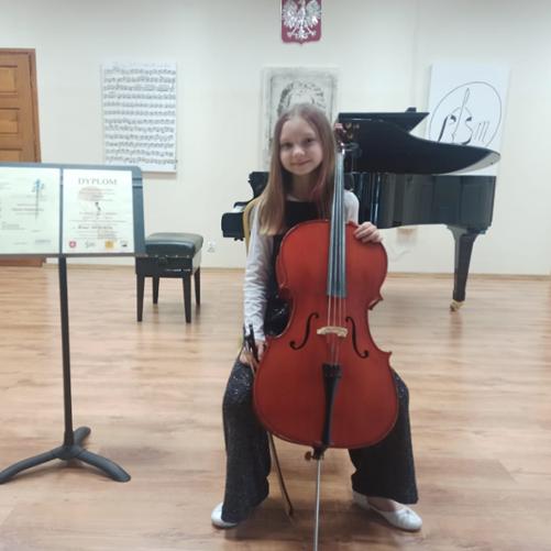 Na zdjęciu siedzi dziewczynka trzymając wiolonczelę obok ustawiono pulpit z Dyplomami w tle z napisami Dyplom Wyróżnienia i Dyplom za zajęcie II miejsca.. W tle fortepian i portrety Jana Sebastiana Bacha, oraz Logo Szkoły i partytura J. S. Bacha.