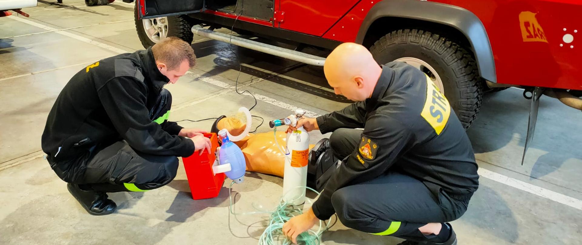 Dwóch strażaków kucających przy manekinie z respiratorem i butlą z tlenem