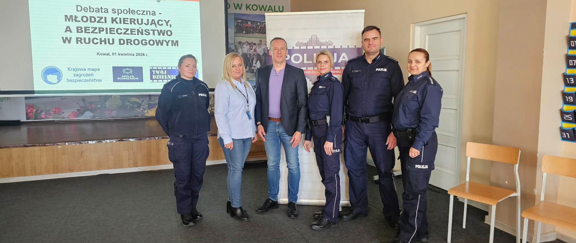 Zdjęcie przedstawia grupę sześciu osób pozujących do wspólnej fotografii po zakończeniu debaty społecznej. Spotkanie odbywa się w sali konferencyjnej, prawdopodobnie w Kowalu. W grupie znajduje się czworo funkcjonariuszy policji w pełnym umundurowaniu oraz dwoje cywilów.
Postacie i układ (od lewej do prawej)
Policjantka: Kobieta o jasnych, spiętych włosach, ubrana w granatowy mundur policyjny.
Kobieta (cywil): Ma długie, blond włosy, ubrana jest w jasnoniebieską koszulę i jeansy.
Mężczyzna (cywil): Wysoki mężczyzna w ciemnej marynarce, fioletowym t-shircie i jeansach. Stoi w centrum grupy.
Druga policjantka: Kobieta o jasnych włosach, w granatowym mundurze.
Policjant: Wysoki, postawny mężczyzna w granatowym mundurze policyjnym.
Trzecia policjantka: Kobieta o ciemnych włosach spiętych w kucyk, również w pełnym umundurowaniu.
Tło i ważne detale
Ekran projekcyjny: Po lewej stronie, w głębi, widać duży ekran z wyświetloną prezentacją. Napis na slajdzie głosi: „Debata społeczna – MŁODZI KIERUJĄCY, A BEZPIECZEŃSTWO W RUCHU DROGOWYM. Kowal, 01 kwietnia 2026 r.”. Na dole slajdu widoczne są logotypy akcji: „Krajowa mapa zagrożeń bezpieczeństwa”, „Moja Komenda” oraz „Twój Dzielnicowy”.
Banner: Po prawej stronie za grupą stoi biało-fioletowy roll-up z dużym napisem „POLICJA” oraz logo Komendy Powiatowej Policji.
Sala: Podłoga wyłożona jest ciemną wykładziną, a w tle widać fragment podestu lub sceny.
Atmosfera
Zdjęcie ma charakter oficjalny i dokumentuje współpracę policji z lokalną społecznością. Wszyscy uczestnicy patrzą w stronę obiektywu z poważnymi, ale pogodnymi minami.