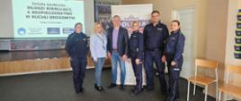 Zdjęcie przedstawia grupę sześciu osób pozujących do wspólnej fotografii po zakończeniu debaty społecznej. Spotkanie odbywa się w sali konferencyjnej, prawdopodobnie w Kowalu. W grupie znajduje się czworo funkcjonariuszy policji w pełnym umundurowaniu oraz dwoje cywilów.
Postacie i układ (od lewej do prawej)
Policjantka: Kobieta o jasnych, spiętych włosach, ubrana w granatowy mundur policyjny.
Kobieta (cywil): Ma długie, blond włosy, ubrana jest w jasnoniebieską koszulę i jeansy.
Mężczyzna (cywil): Wysoki mężczyzna w ciemnej marynarce, fioletowym t-shircie i jeansach. Stoi w centrum grupy.
Druga policjantka: Kobieta o jasnych włosach, w granatowym mundurze.
Policjant: Wysoki, postawny mężczyzna w granatowym mundurze policyjnym.
Trzecia policjantka: Kobieta o ciemnych włosach spiętych w kucyk, również w pełnym umundurowaniu.
Tło i ważne detale
Ekran projekcyjny: Po lewej stronie, w głębi, widać duży ekran z wyświetloną prezentacją. Napis na slajdzie głosi: „Debata społeczna – MŁODZI KIERUJĄCY, A BEZPIECZEŃSTWO W RUCHU DROGOWYM. Kowal, 01 kwietnia 2026 r.”. Na dole slajdu widoczne są logotypy akcji: „Krajowa mapa zagrożeń bezpieczeństwa”, „Moja Komenda” oraz „Twój Dzielnicowy”.
Banner: Po prawej stronie za grupą stoi biało-fioletowy roll-up z dużym napisem „POLICJA” oraz logo Komendy Powiatowej Policji.
Sala: Podłoga wyłożona jest ciemną wykładziną, a w tle widać fragment podestu lub sceny.
Atmosfera
Zdjęcie ma charakter oficjalny i dokumentuje współpracę policji z lokalną społecznością. Wszyscy uczestnicy patrzą w stronę obiektywu z poważnymi, ale pogodnymi minami.