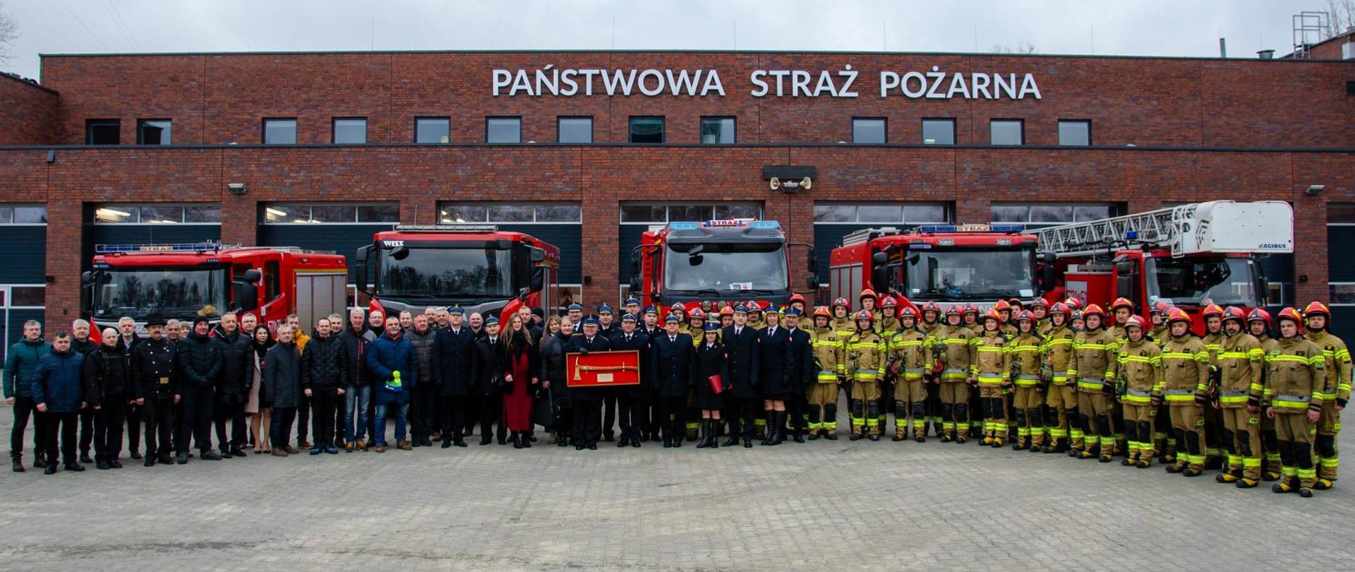 Grupa strażaków w umundurowaniu bojowym oraz funkcjonariuszy i gości cywilnych stoi przed budynkiem z napisem „Państwowa Straż Pożarna”. W tle widoczne są dwa czerwone wozy strażackie zaparkowane przed garażami jednostki.