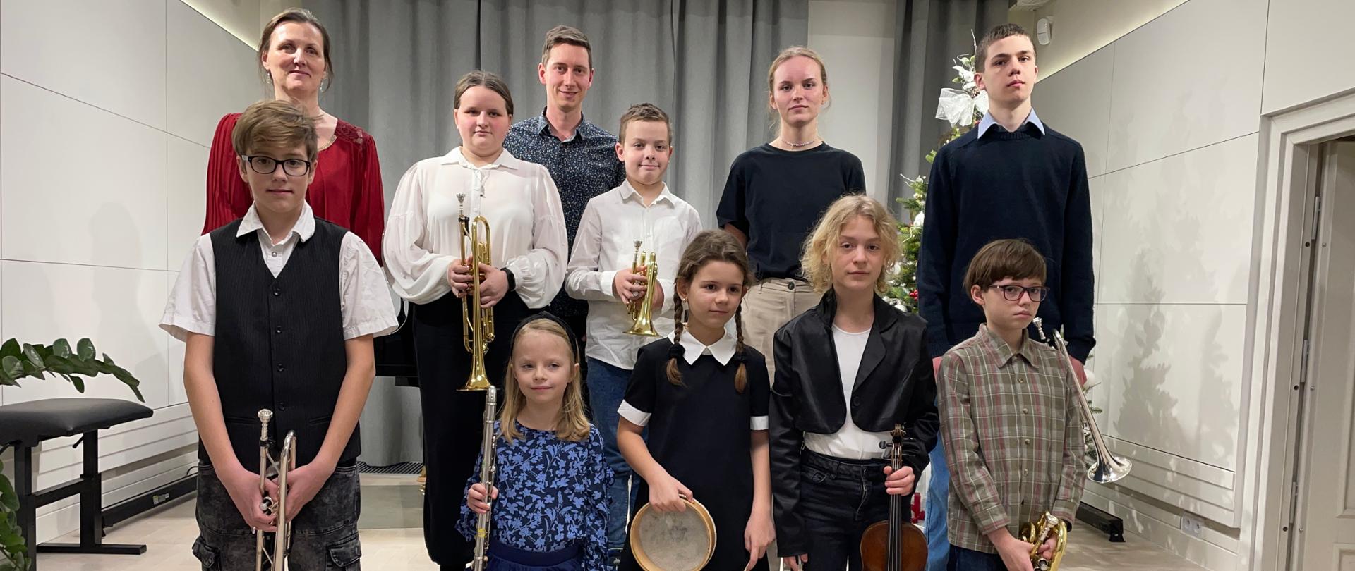 Na zdjęciu znajdują się uczniowie ustawieni w dwurzędzie na stopniach sceny. Z lewe strony w drugim rzędzie stoi pani akompaniator, a na środku z uczniami nauczyciel klasy trąbki. Uczniowie trzymają w dłoniach instrumenty: trąbki, flet, bębenek i skrzypce. Z pierwszym uczniem od prawej widać fragment choinki. Tłem do zdjęcia są zasunięte częściowo szare kotary. Pozostałe tło jest białe. 