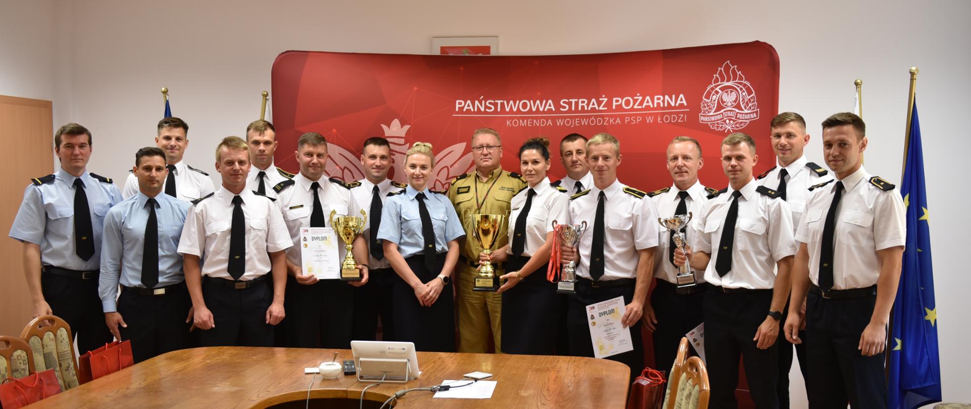 na zdjęciu grupa strażaków stojąca w sali, pozują do zdjęcia, prezentują cztery zdobyte puchary, za nimi ścianka z napisem państwowa straż pożarna