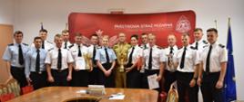 na zdjęciu grupa strażaków stojąca w sali, pozują do zdjęcia, prezentują cztery zdobyte puchary, za nimi ścianka z napisem państwowa straż pożarna