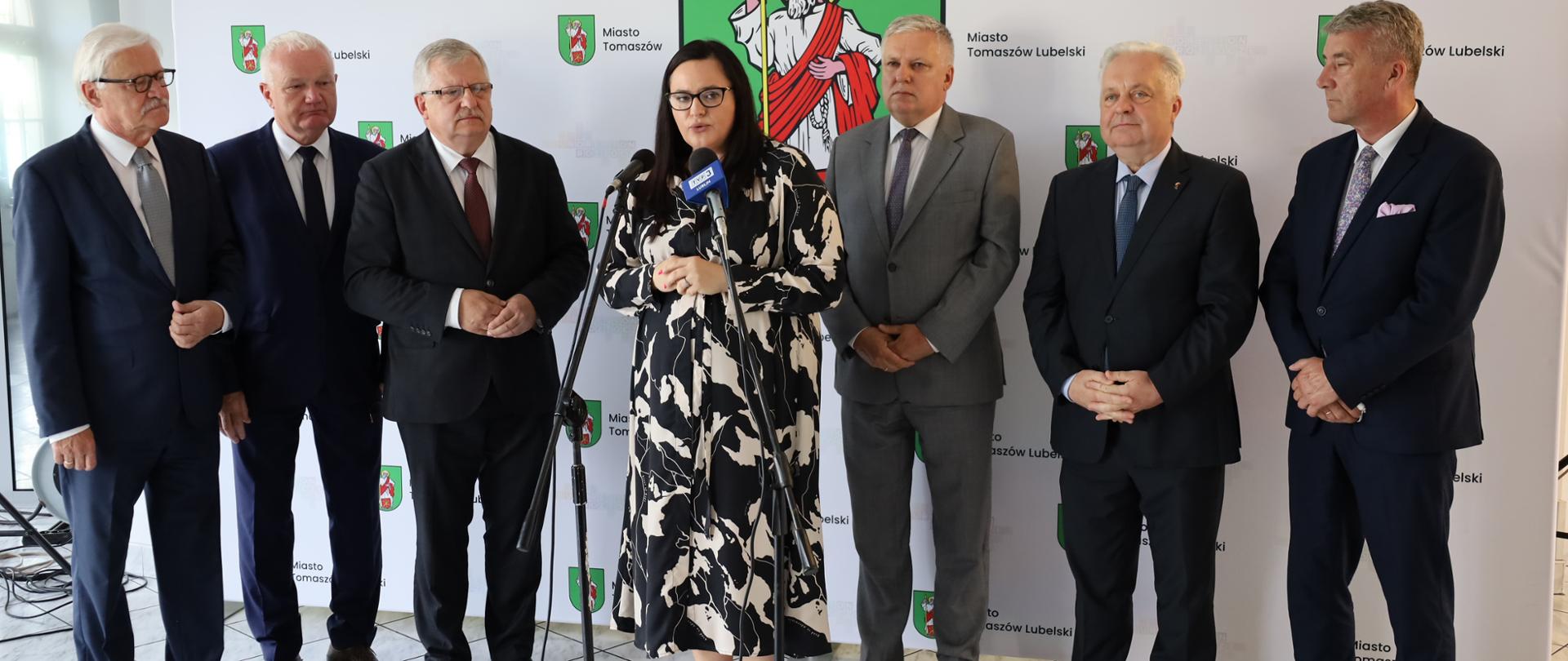 Podczas konferencji prasowej wiceminister Małgorzata Jarosińska-Jedynak mówi do ustawionych mikrofonów. Za nią grupa mężczyzn. Z tyłu ścianka miasta Tomaszów Lubelski.