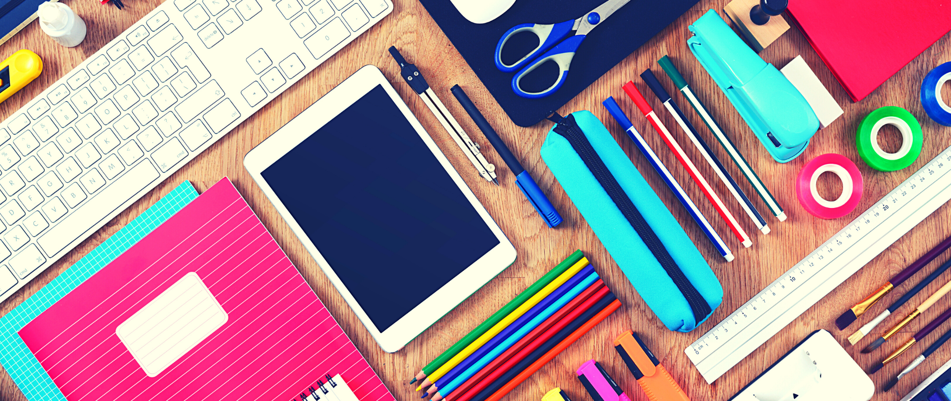 Biurko, na blacie pośrodku biały tablet, dookoła różnokolorowe przybory szkolne - notatnik, kredki, pędzelki itp. 