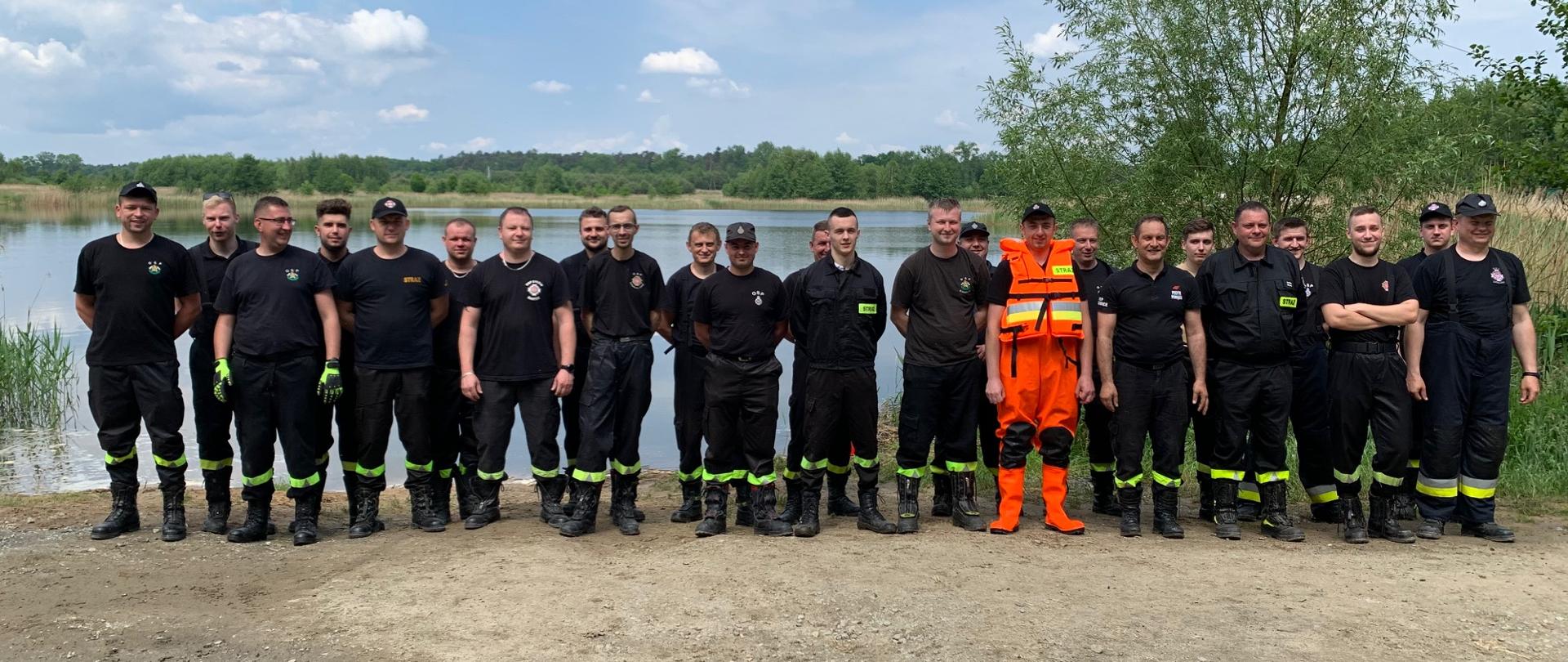 Na zdjęciu grupa strażaków w umundurowaniu czarnym (jedna osoba w pomarańczowym stroju do działania na wodzie). Strażacy stoją w dwuszeregu, w tle widoczny zalew wodny. 