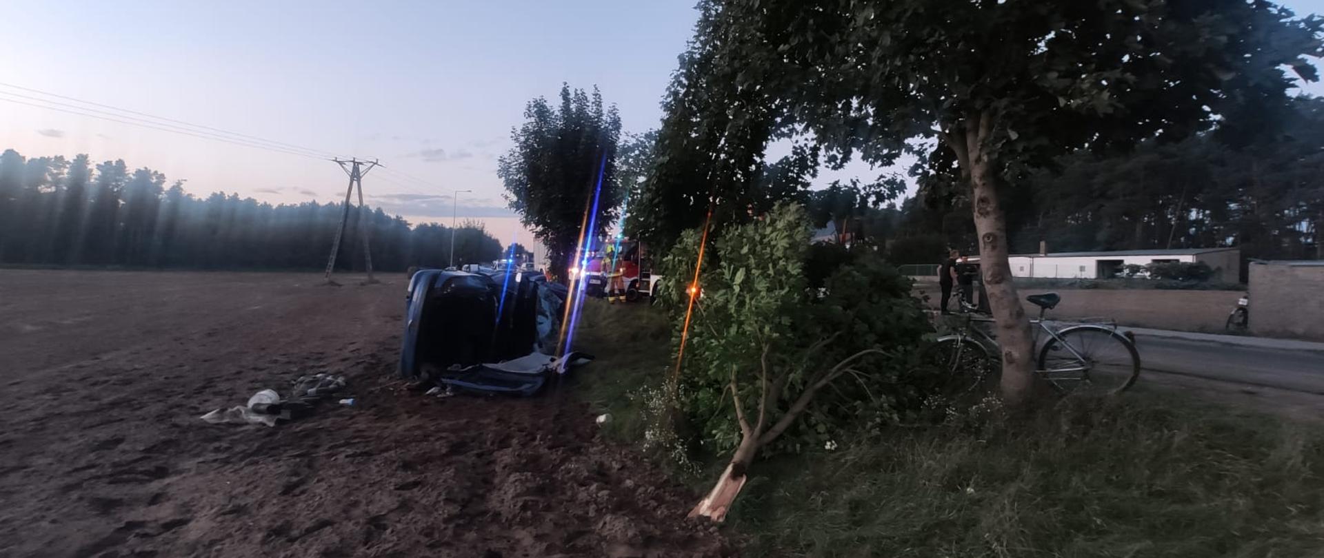 Zdjęcie przedstawia samochód osobowy, który po wypadnięciu z drogi leży na boku. Przy pomocy narzędzi hydraulicznych został odcięty dach samochodu. Zdjęcie zrobione na poboczu drogi, obok rosna drzewa.