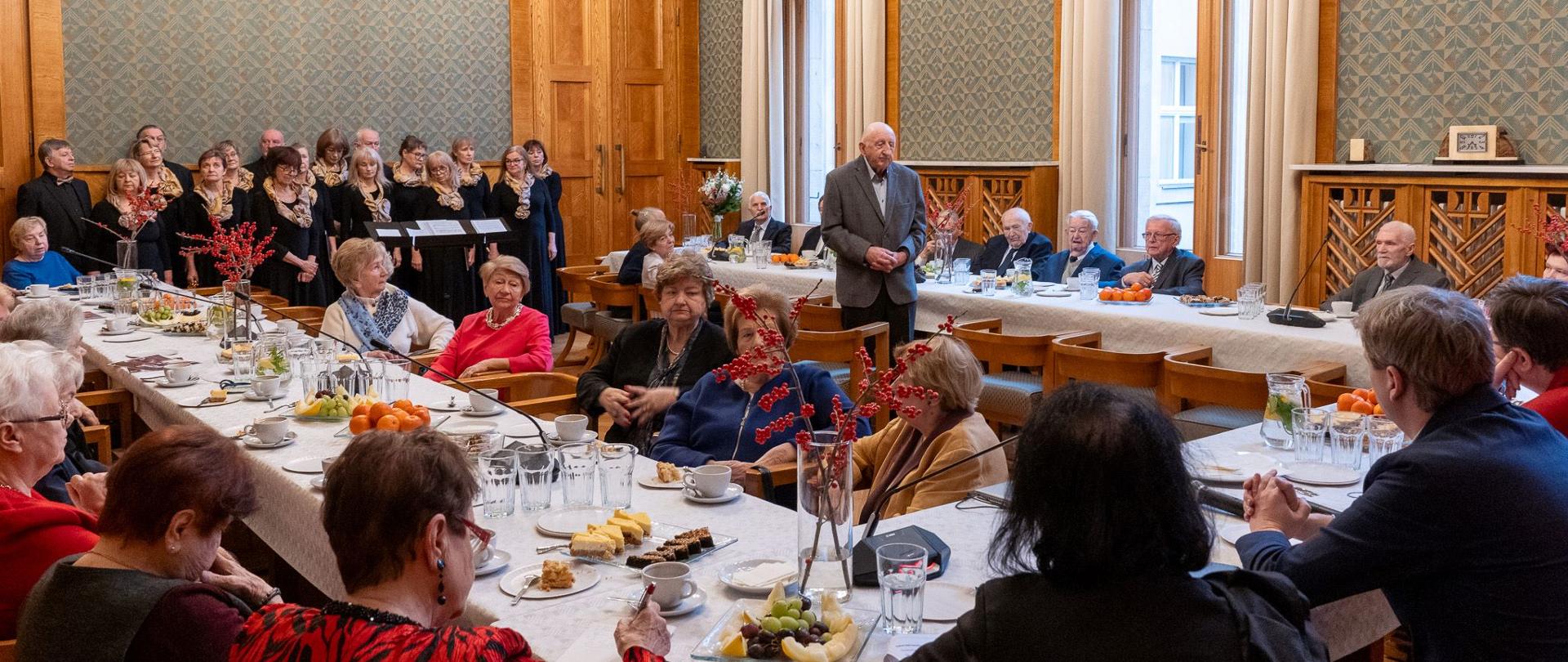 Duża sala ze ścianami w zielony wzorek, przy drewnianych stołach ustawionych w czworokąt siedzi dużo elegancko ubranych osób. Pod ścianą stoi chór.