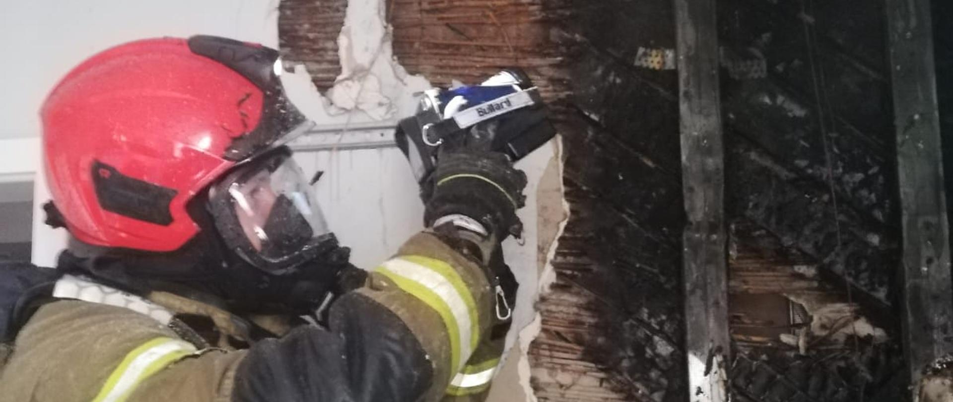 Zdjęcie przedstawia strażaka, który sprawdza kamerą termowizyjną gdzie znajdują się miejsca o podwyższonej temperaturze. Strażak ubrany jest w ubranie bojowe oraz hełm. Strażak ma na sobie środki ochrony osobistej apart oddechowy oraz maskę. 